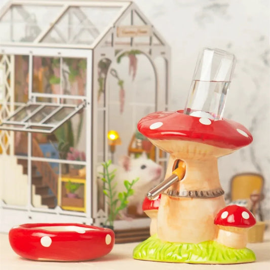 A mushroom-shaped bird feeder and waterer set, featuring a large red and white spotted mushroom cap with a clear plastic water bottle inserted, a smaller red mushroom at the base, a red dish, and a small green grassy base. A small white hamster is visible behind it inside a miniature glass terrarium.