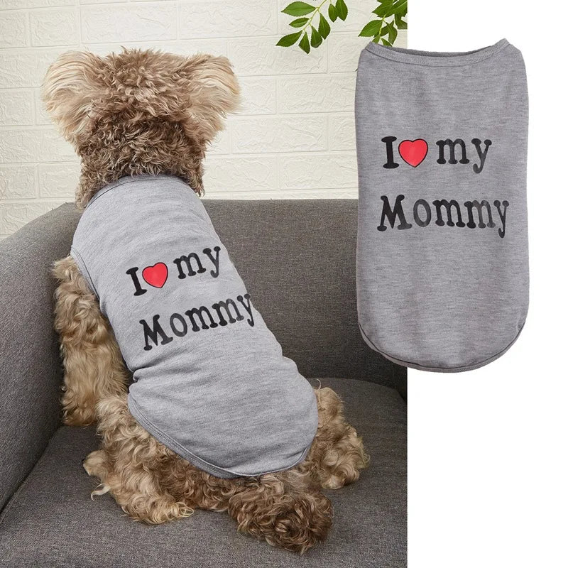 A small fluffy dog sits on a gray sofa wearing a light gray pet shirt printed with 'I [heart] my Mommy' in black text. A second flat view of the same shirt appears on the right against a white background.