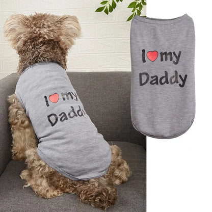 A small curly-haired dog sits on a gray couch wearing a heather gray pet shirt with the printed phrase 'I ♥ my Daddy' in black text with a red heart. A second flat view of the same shirt appears on the right against a white background.