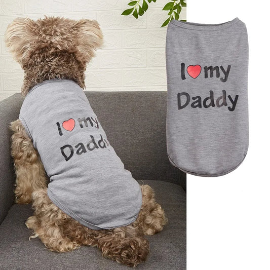 A small curly-haired dog sits on a gray couch wearing a heather gray pet shirt with the printed phrase 'I ♥ my Daddy' in black text with a red heart. A second flat view of the same shirt appears on the right against a white background.
