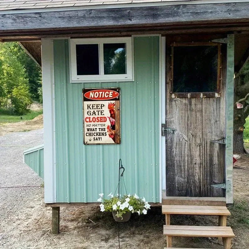 A rustic wooden chicken coop with light green vertical siding, a weathered dark wood door, a small window, and a metal sign that reads 'KEEP GATE CLOSED'. A small hanging flower basket sits in front of the door on a gravel surface outdoors.