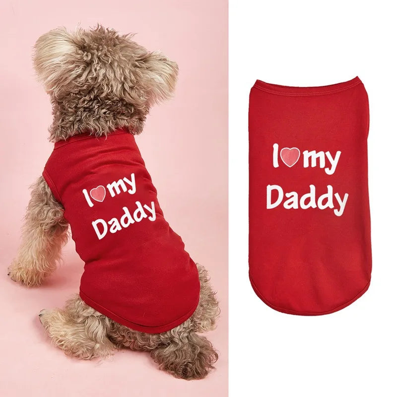 A small fluffy dog wearing a red pet shirt with the white text 'I love my Daddy' and a heart symbol, shown on the left against a pink background. On the right, the same red pet shirt is shown flat on a white background with the same text.
