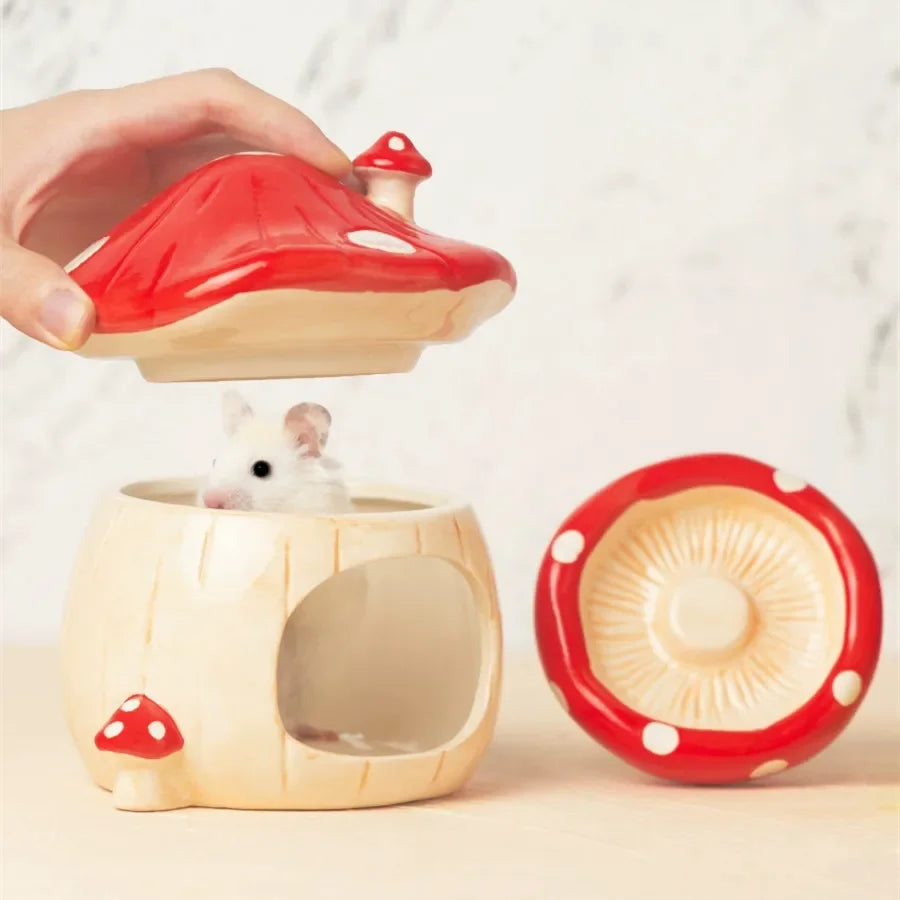 A ceramic hamster hide shaped like a mushroom, with a small white hamster visible inside. A human hand is lifting the red-and-cream mushroom lid, and a separate lid piece sits to the right on a light neutral background.