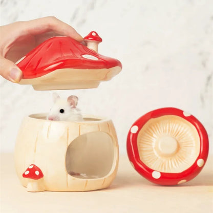 A ceramic hamster hide shaped like a mushroom, with a small white hamster visible inside. A human hand is lifting the red-and-cream mushroom lid, and a separate lid piece sits to the right on a light neutral background.