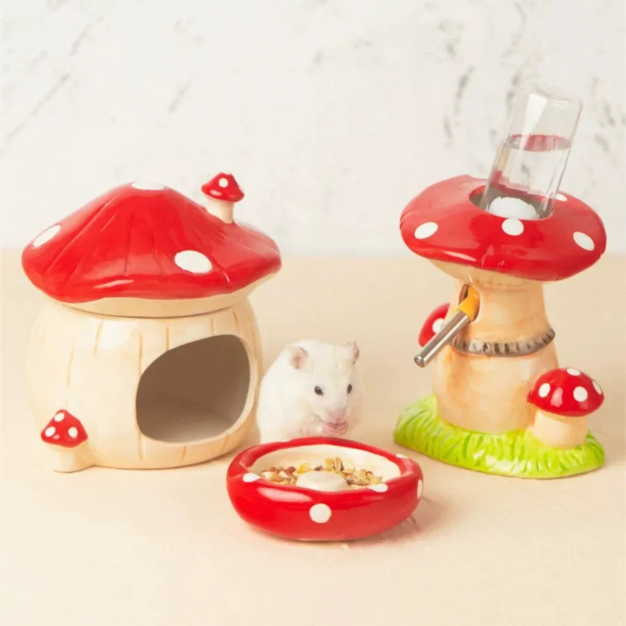 A small white hamster sits between two mushroom-shaped pet accessories on a light beige surface against a pale marble-like background. One mushroom house has a red cap with white spots and an open front; the other is a drinking fountain with a red cap, green base, and clear water bottle.