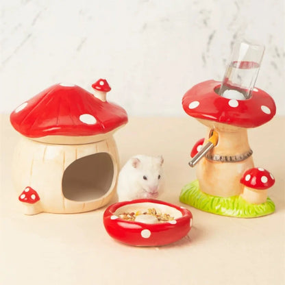 A small white hamster sits between two mushroom-shaped pet accessories on a light beige surface against a pale marble-like background. One mushroom house has a red cap with white spots and an open front; the other is a drinking fountain with a red cap, green base, and clear water bottle.