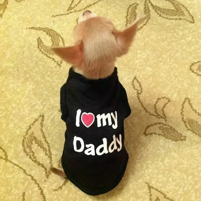A small dog seen from behind wearing a black pet shirt with the text 'I [heart] my Daddy' printed on the back, sitting on a beige patterned carpet.