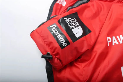 A close-up of a red puffer jacket featuring a black Supreme box logo patch, a black The Face of the North logo patch, and white lettering on a white background.