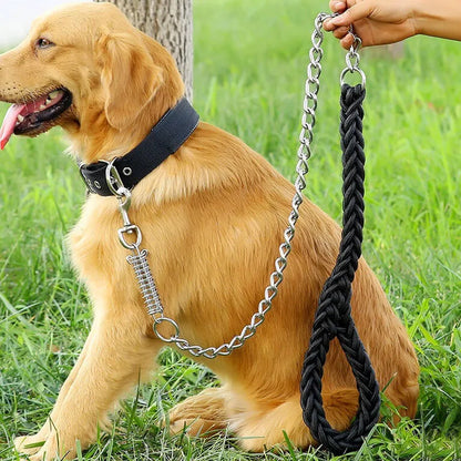 A golden retriever dog wearing a black collar with a heavy-duty black braided leash attached by a silver chain. A human hand is holding the end of the leash, set against a green grass background.