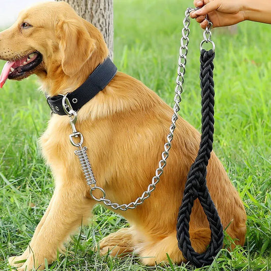 A golden retriever dog wearing a black collar with a heavy-duty black braided leash attached by a silver chain. A human hand is holding the end of the leash, set against a green grass background.