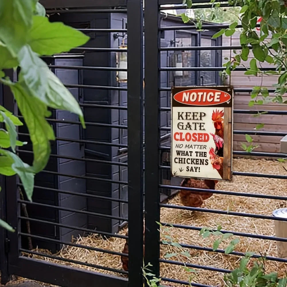 A black metal chicken coop gate with a sign that reads 'NOTICE KEEP GATE CLOSED NO MATTER WHAT THE CHICKENS SAY!' in a garden setting with straw bedding and chickens visible inside.