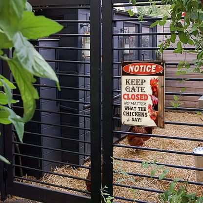 A black metal chicken coop gate with a sign that reads 'NOTICE KEEP GATE CLOSED NO MATTER WHAT THE CHICKENS SAY!' in a garden setting with straw bedding and chickens visible inside.
