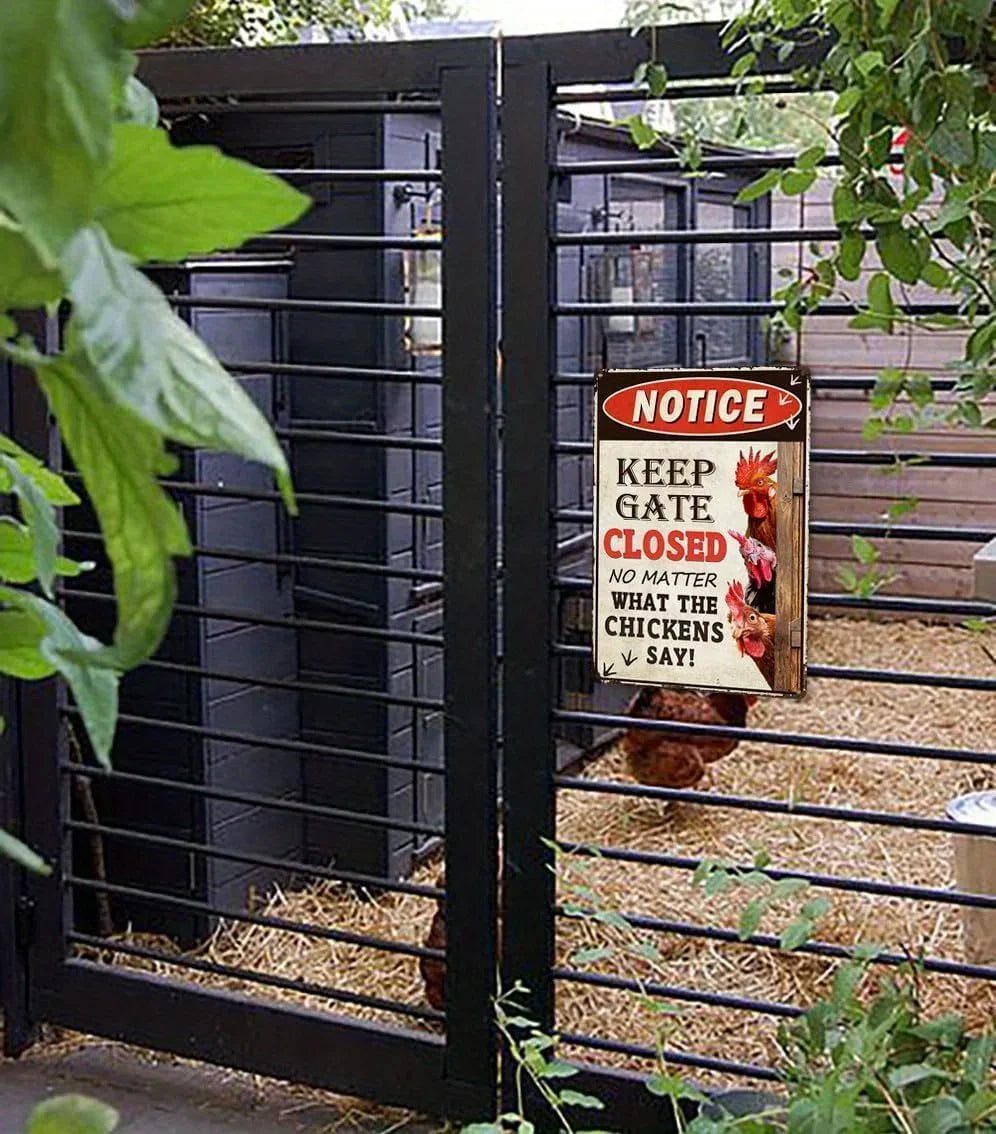 A black metal chicken coop gate with a hanging 'Notice' sign that reads 'Keep Gate Closed No Matter What the Chickens Say!' in a garden setting with straw bedding and a chicken visible inside.