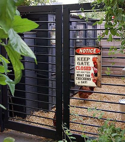 A black metal chicken coop gate with a hanging 'Notice' sign that reads 'Keep Gate Closed No Matter What the Chickens Say!' in a garden setting with straw bedding and a chicken visible inside.