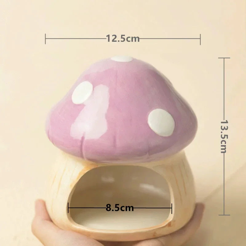 A mushroom-shaped ceramic pet house with a purple cap, white spots, and a beige wooden base, held in a hand. Black dimension lines and text overlay show measurements of 12.