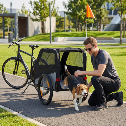 A man kneels beside a black cargo trailer attached to a bicycle on a paved path in a park, with a small brown and white dog standing in the trailer. The scene is outdoors with grass, trees, and a bright orange flag visible in the background.