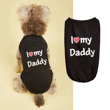 A small fluffy dog wearing a black pet shirt with the text 'I [heart] my Daddy' in white, shown from the back against a pale yellow background. A second flat view of the same black shirt appears on the right with the same slogan and a pink heart graphic.